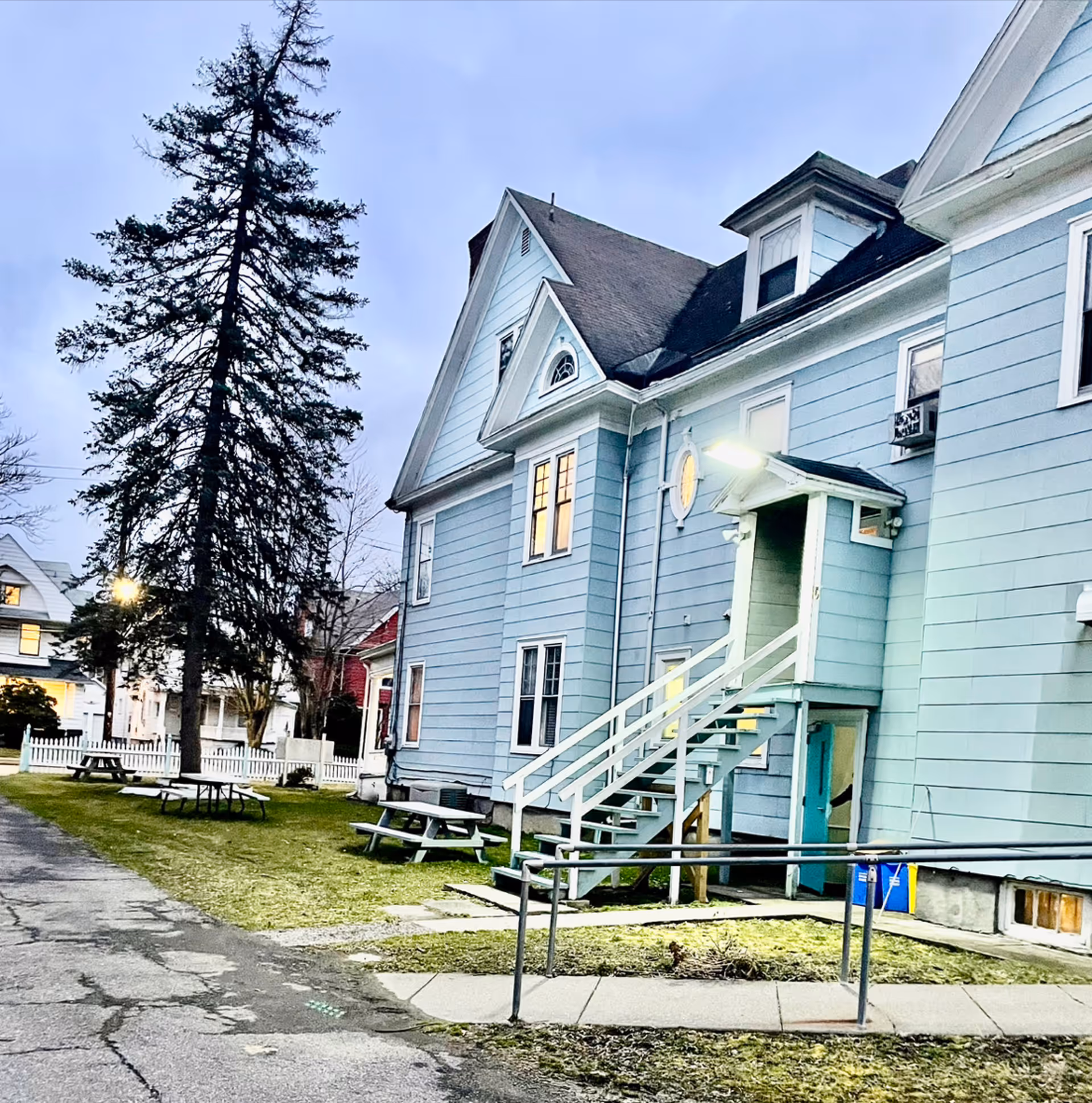 Light-blue multi-story house exterior with outdoor stairs, a wheelchair ramp, picnic tables on the lawn, and a tall evergreen tree at dusk.