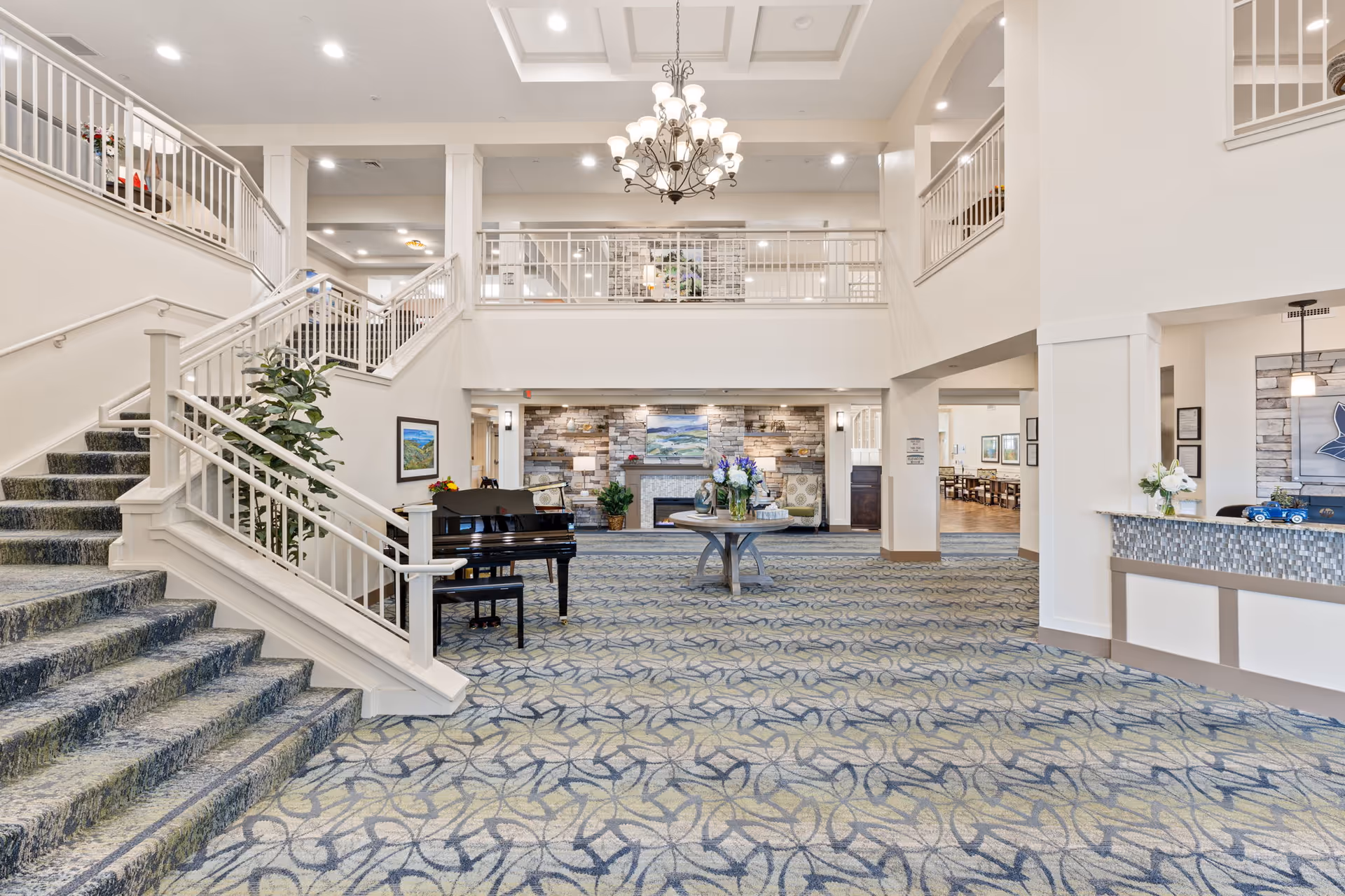 Spacious and well-lit senior living facility lobby with a patterned carpet, a grand piano, a round table with flower arrangements, a stone fireplace, and a chandelier hanging from the ceiling. There is a staircase on the left side leading to an upper floor with railings, and a reception desk on the right side.