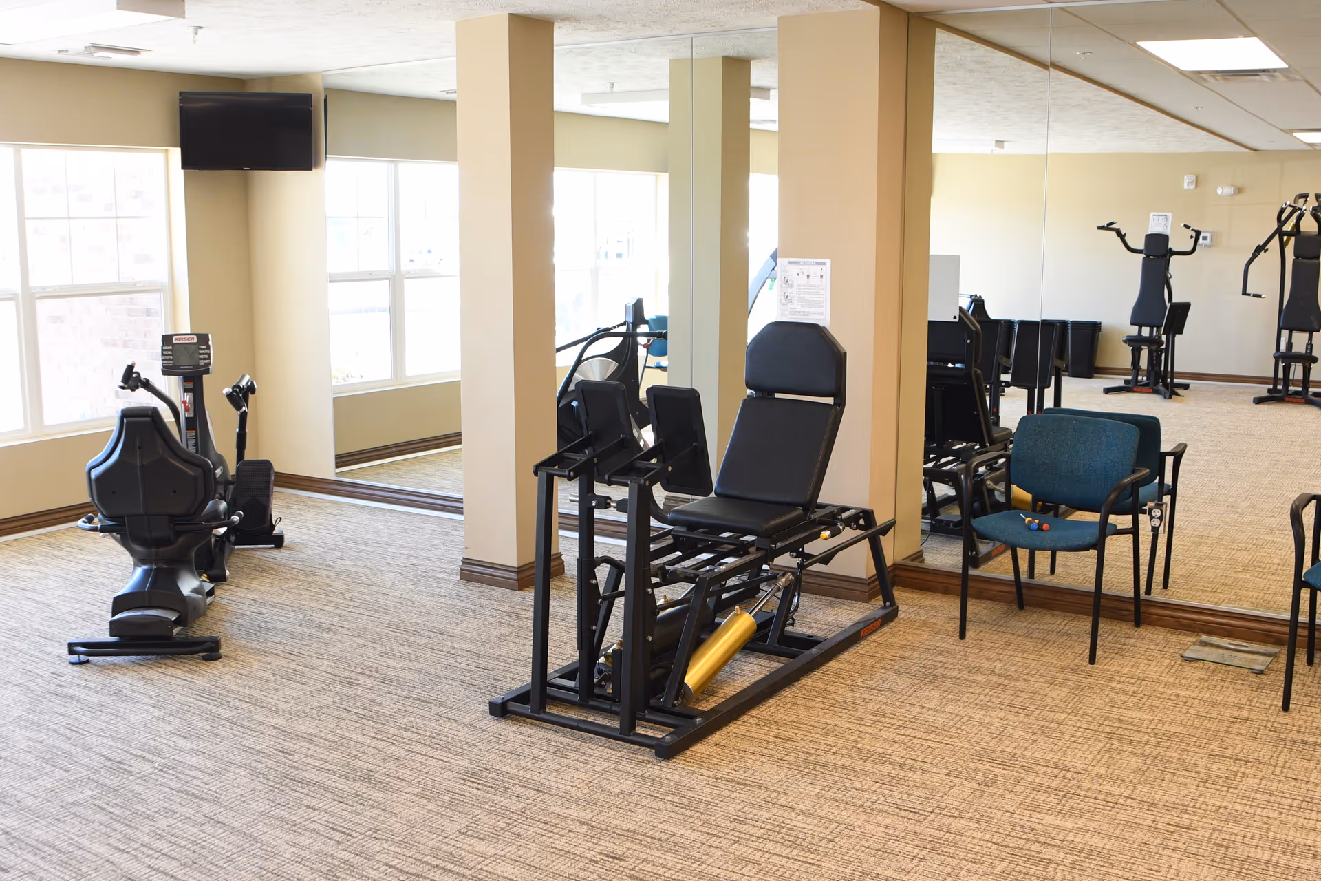 A bright exercise room with weight machines, a recumbent bike, chairs, and mirrors along one wall.
