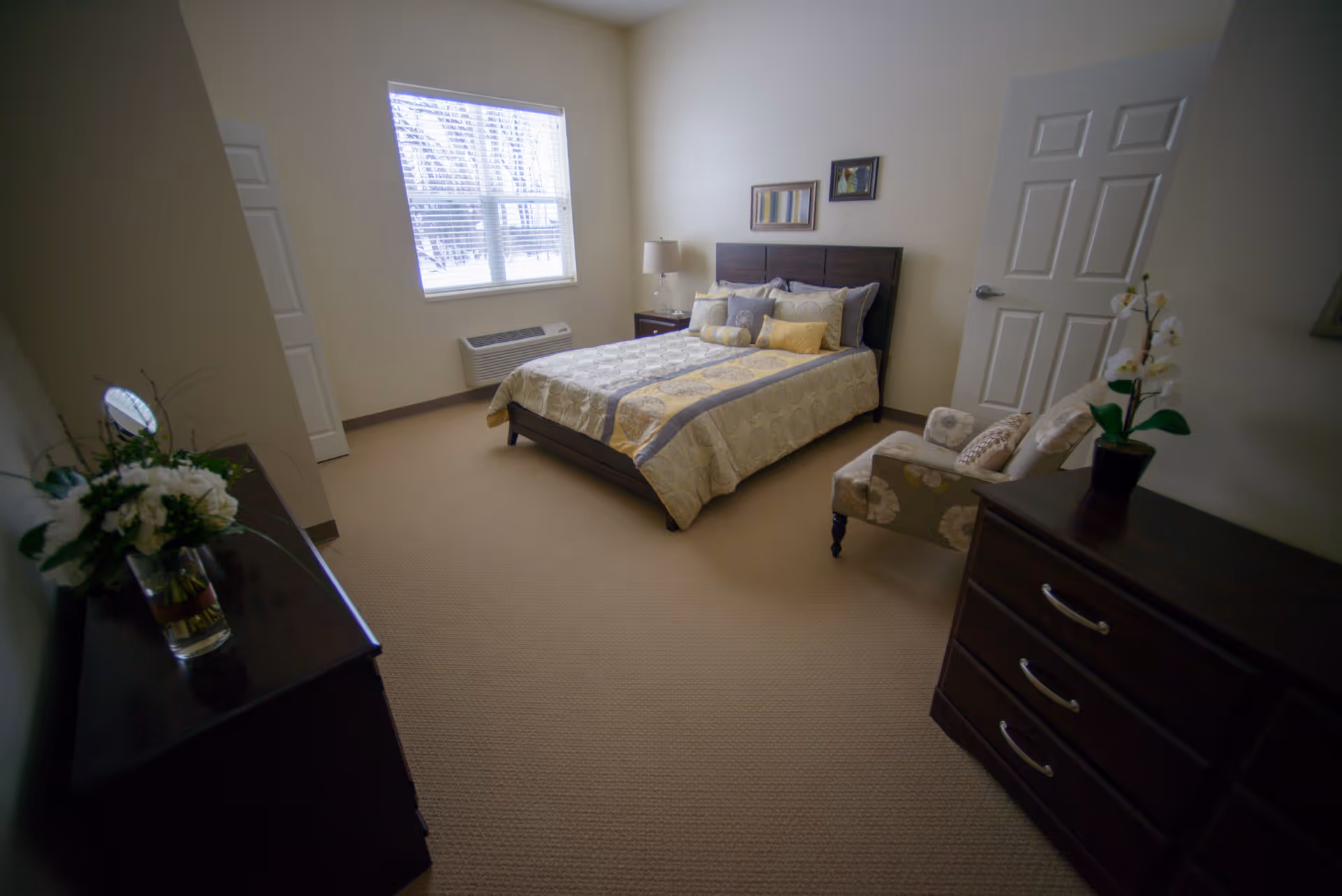 A neatly arranged bedroom with a bed covered in a beige and yellow patterned comforter, several pillows, a nightstand with a lamp, a floral armchair, a dresser with a potted orchid, and a window letting in natural light.