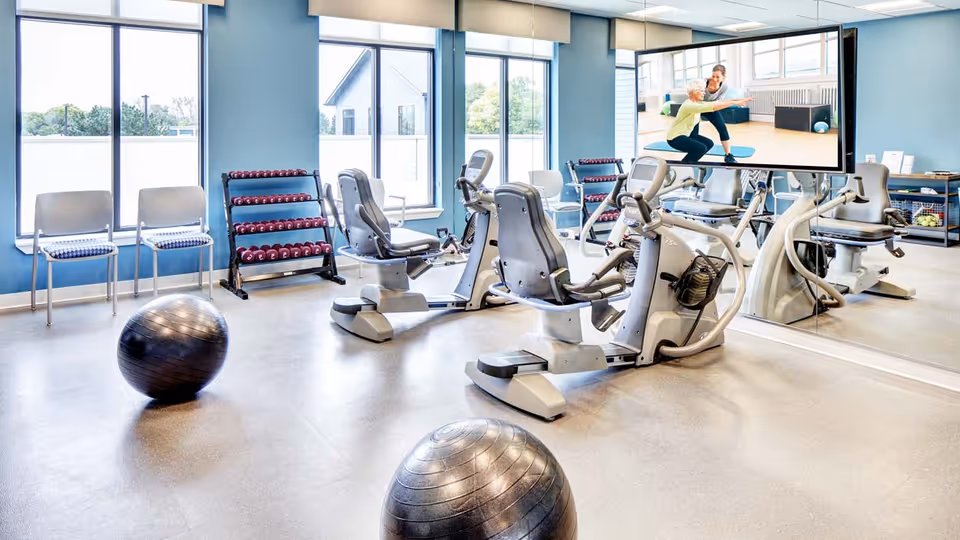 A bright fitness room with exercise equipment including stationary recumbent bikes, two large exercise balls on the floor, a rack of dumbbells, and two chairs by the window. A large mirror covers one wall, reflecting the room and a mounted TV screen showing two women exercising.