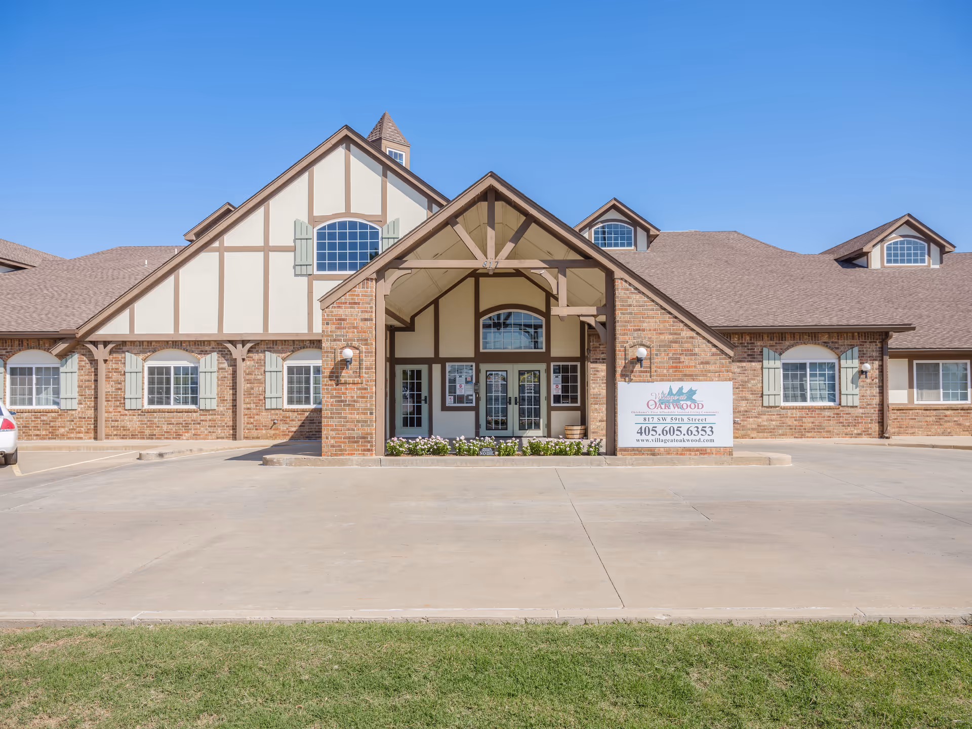 Front exterior view of Village At Oakwood senior living facility with a brick and beige facade, multiple windows with green shutters, a peaked roof, and a covered entrance. There is a sign near the entrance displaying the facility's name, address, phone number, and website.