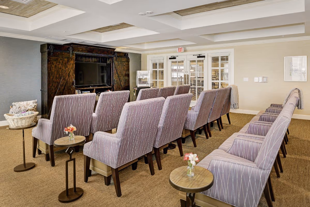 Interior seating room with rows of upholstered chairs facing a large TV cabinet and small side tables with flowers.