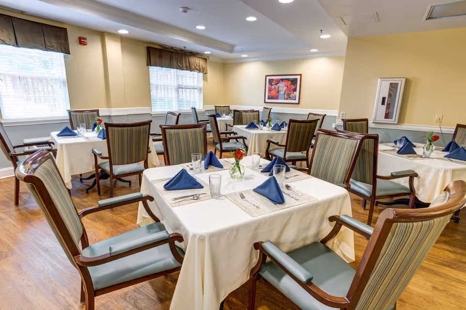 A dining room with several tables covered in white tablecloths, each set with blue folded napkins, glasses, silverware, and small vases with red flowers. The room has wooden floors, striped cushioned chairs, two windows with brown valances, and a framed colorful artwork on the wall.