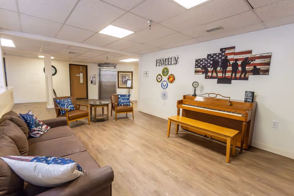 A cozy common area in a senior living facility featuring a brown couch with patriotic-themed pillows, two brown armchairs with blue cushions, a round glass-top table, and a wooden piano with a matching bench. The wall behind the piano is decorated with a large American flag artwork depicting soldiers, a clock, a small sign, and military branch emblems arranged around the word 'FAMILY'. The room has light wood flooring and a white ceiling with recessed lighting. An elevator and a door are visible in the background.
