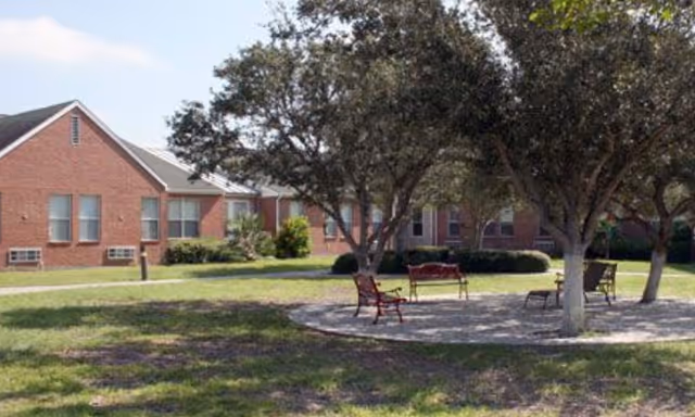 A brick single-story building with a shaded outdoor courtyard of trees and benches on a grassy lawn.