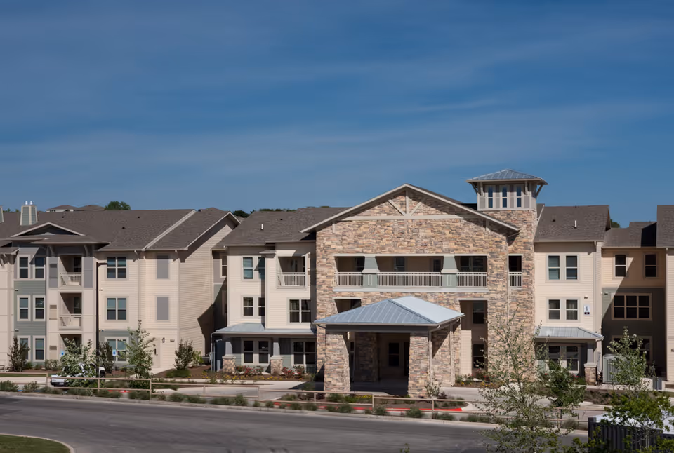 Exterior view of a multi-story senior living facility building with stone and beige siding, balconies, and a covered entrance under a clear blue sky.