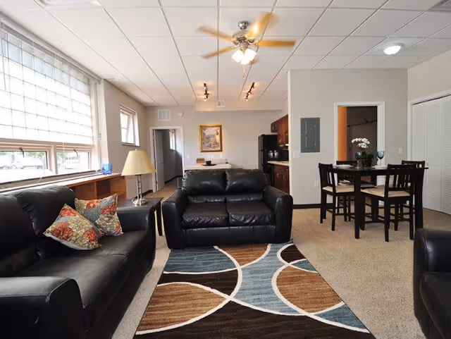 A well-lit living room area with two black leather sofas, a patterned area rug, a ceiling fan with lights, a small dining table with four chairs, and a kitchen area in the background. There is a large window with blinds on the left side and a lamp on a side table next to one of the sofas.