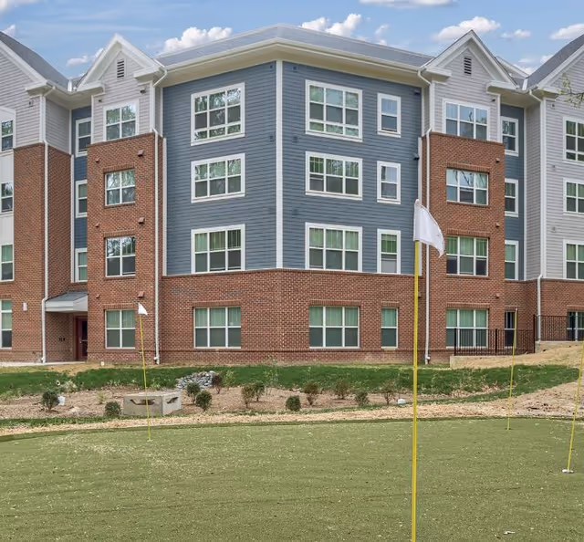Exterior view of a multi-story senior living residence building with a combination of red brick and blue siding. The building has multiple windows and a small putting green with flags in the foreground under a partly cloudy sky.