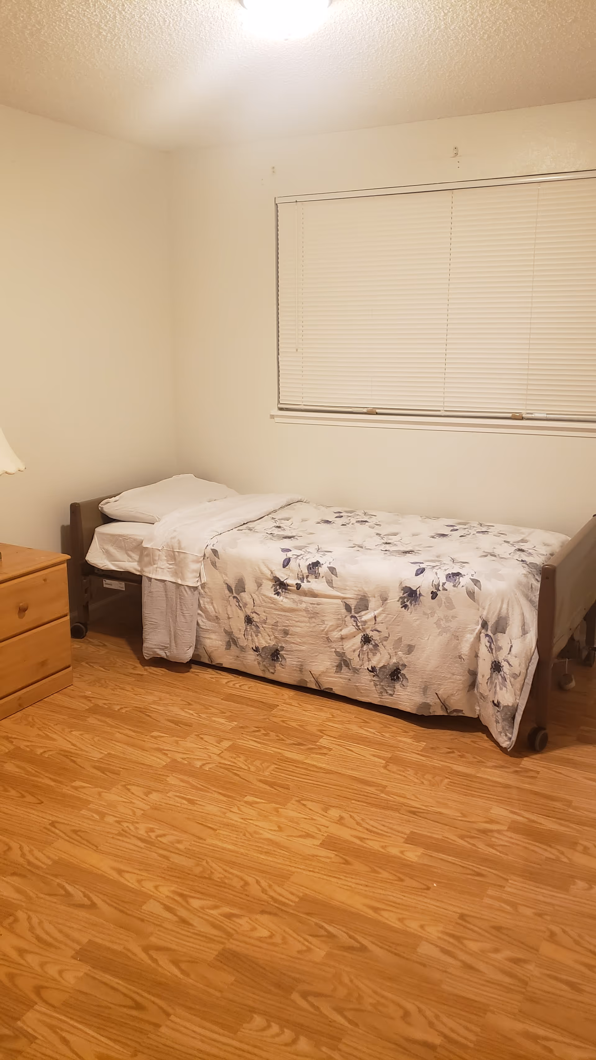 A simple bedroom with a single bed covered with a floral patterned blanket, a wooden nightstand with a lamp, and a window with closed blinds. The floor is wooden and the walls are plain white.