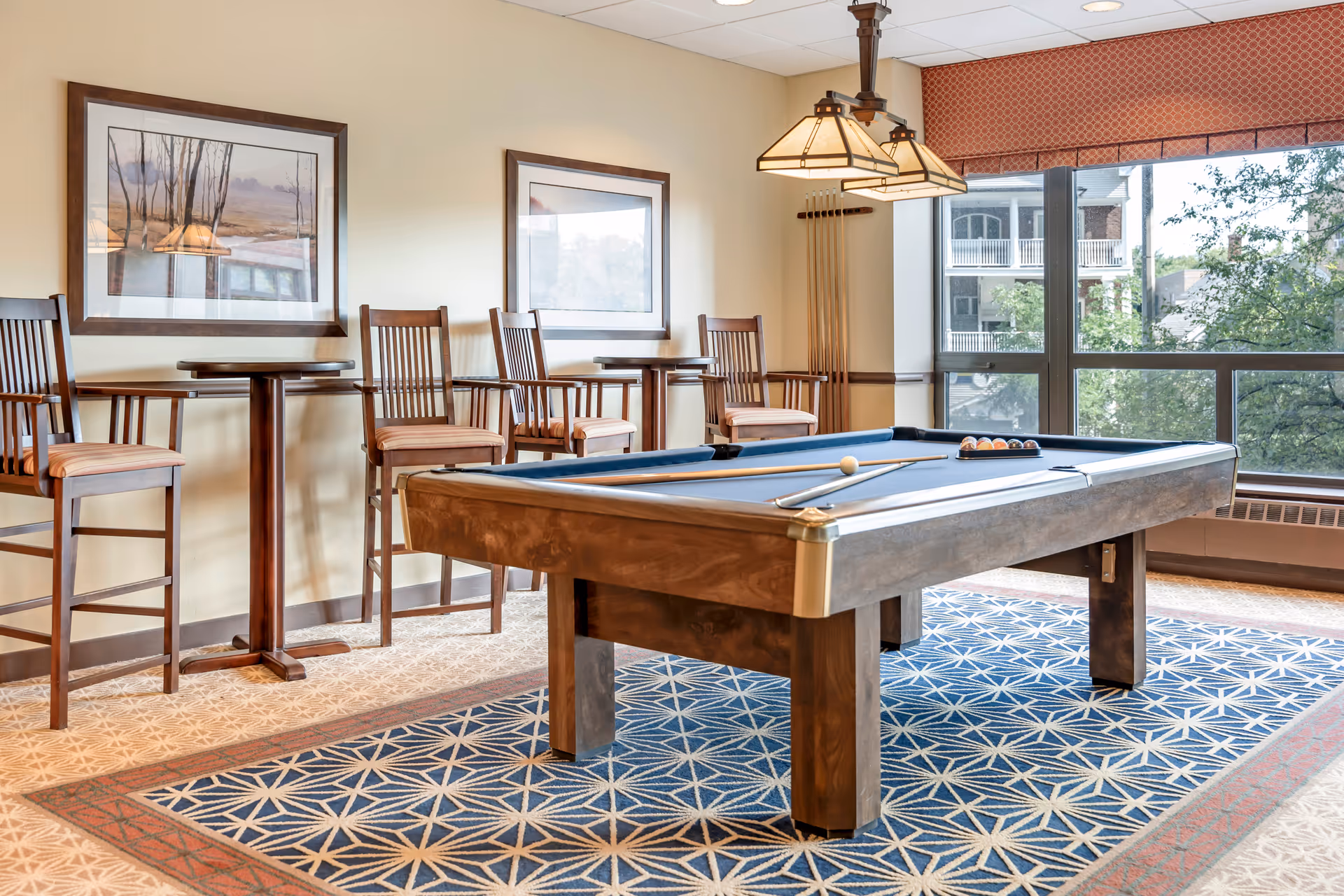 A well-lit game room featuring a pool table with cues and balls arranged on it. Behind the pool table are four wooden chairs with striped cushions and two small round tables. The room has large windows with a view of trees and buildings outside. Two framed pictures hang on the wall, and a patterned carpet covers the floor.
