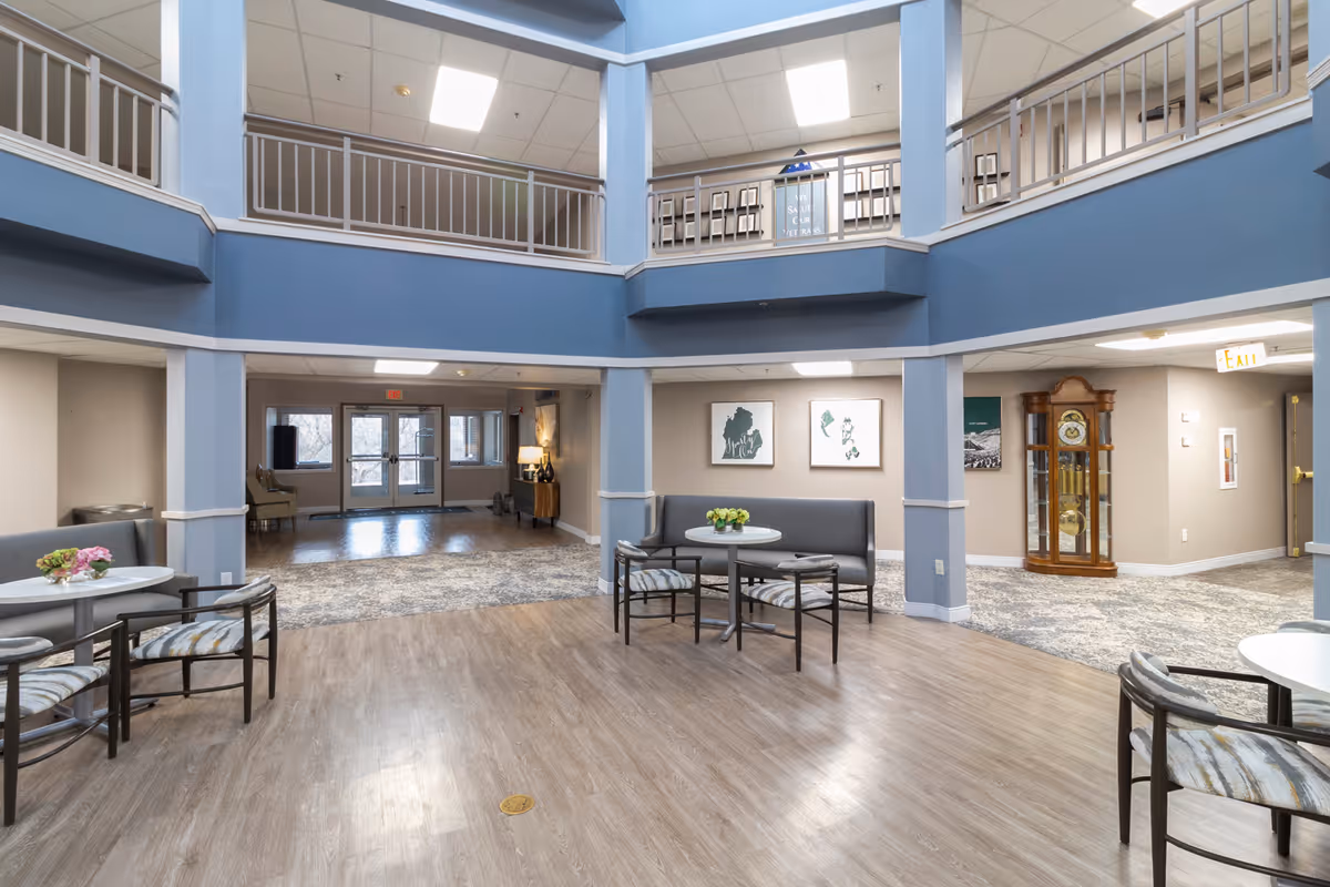 Spacious, well-lit common area in a senior living facility with light blue walls and a two-story open ceiling. The room features small round tables with chairs and built-in bench seating along the walls. There is a grandfather clock in the corner, framed artwork on the walls, and an exit door visible in the background.