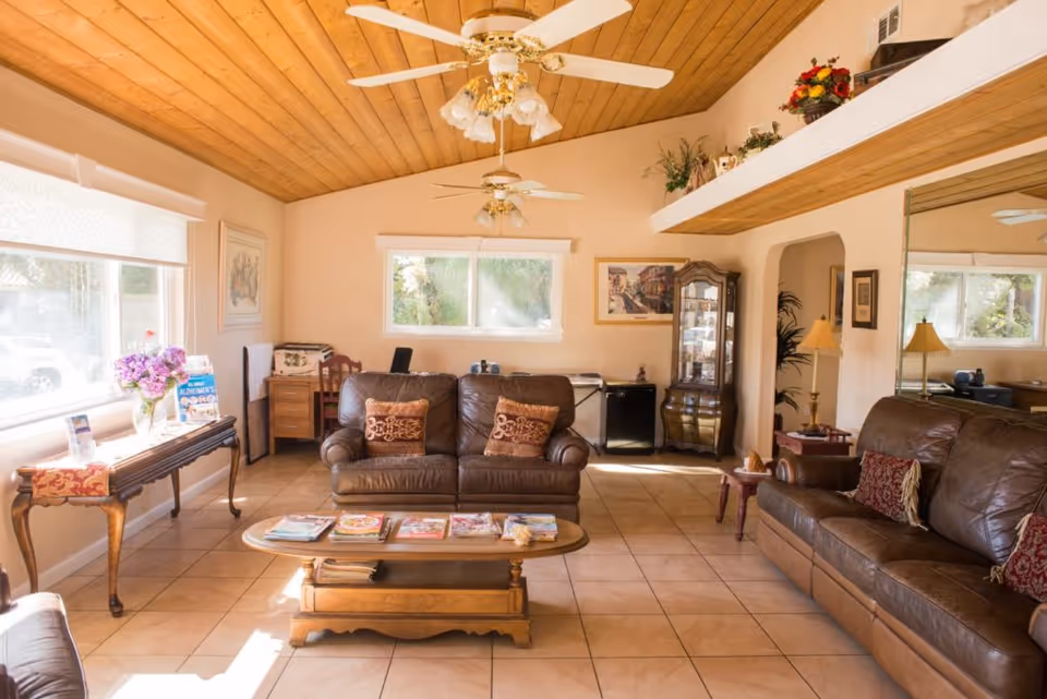 A cozy living room with a wooden ceiling and two ceiling fans. The room features two brown leather sofas with decorative pillows, a wooden coffee table with magazines on it, and a side table with a lamp. There is a large window letting in natural light, a wooden display cabinet, and various framed pictures and plants decorating the space.