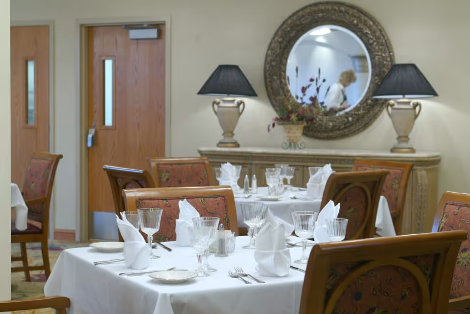 A dining room with tables covered in white tablecloths, set with folded white napkins, glassware, plates, and silverware. The chairs have patterned upholstery. In the background, there is a wooden sideboard with two lamps and a round decorative mirror reflecting a person walking by.