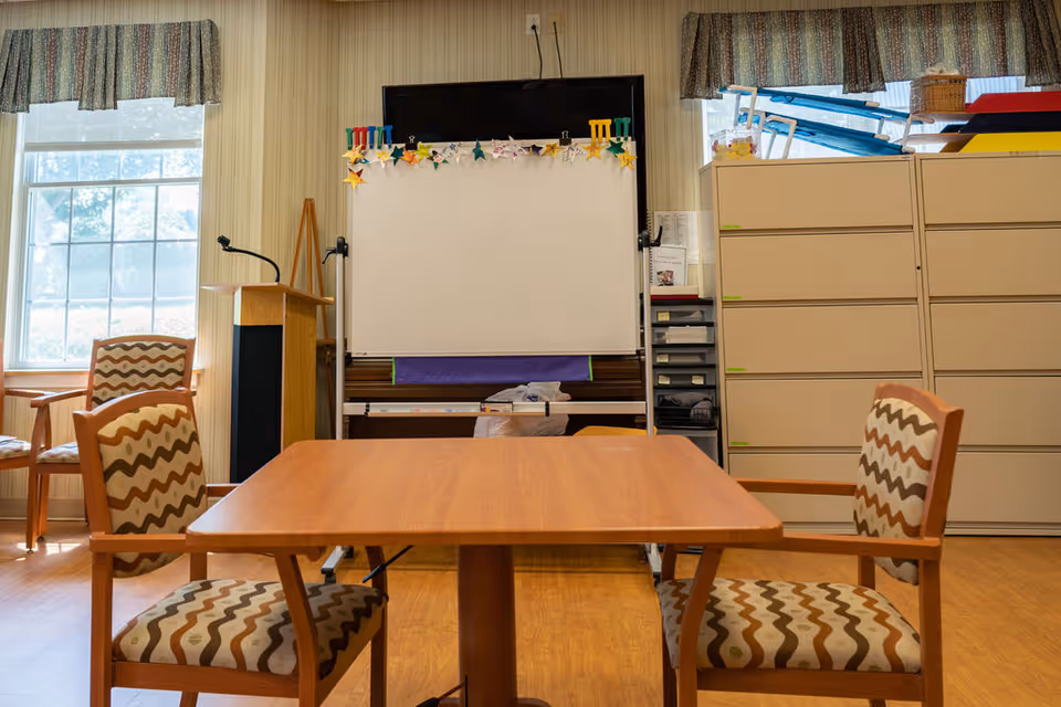 A small meeting or activity room with a wooden table and four patterned chairs. There is a whiteboard on a stand decorated with paper stars, a podium with a microphone, filing cabinets, and windows with patterned valances letting in natural light.