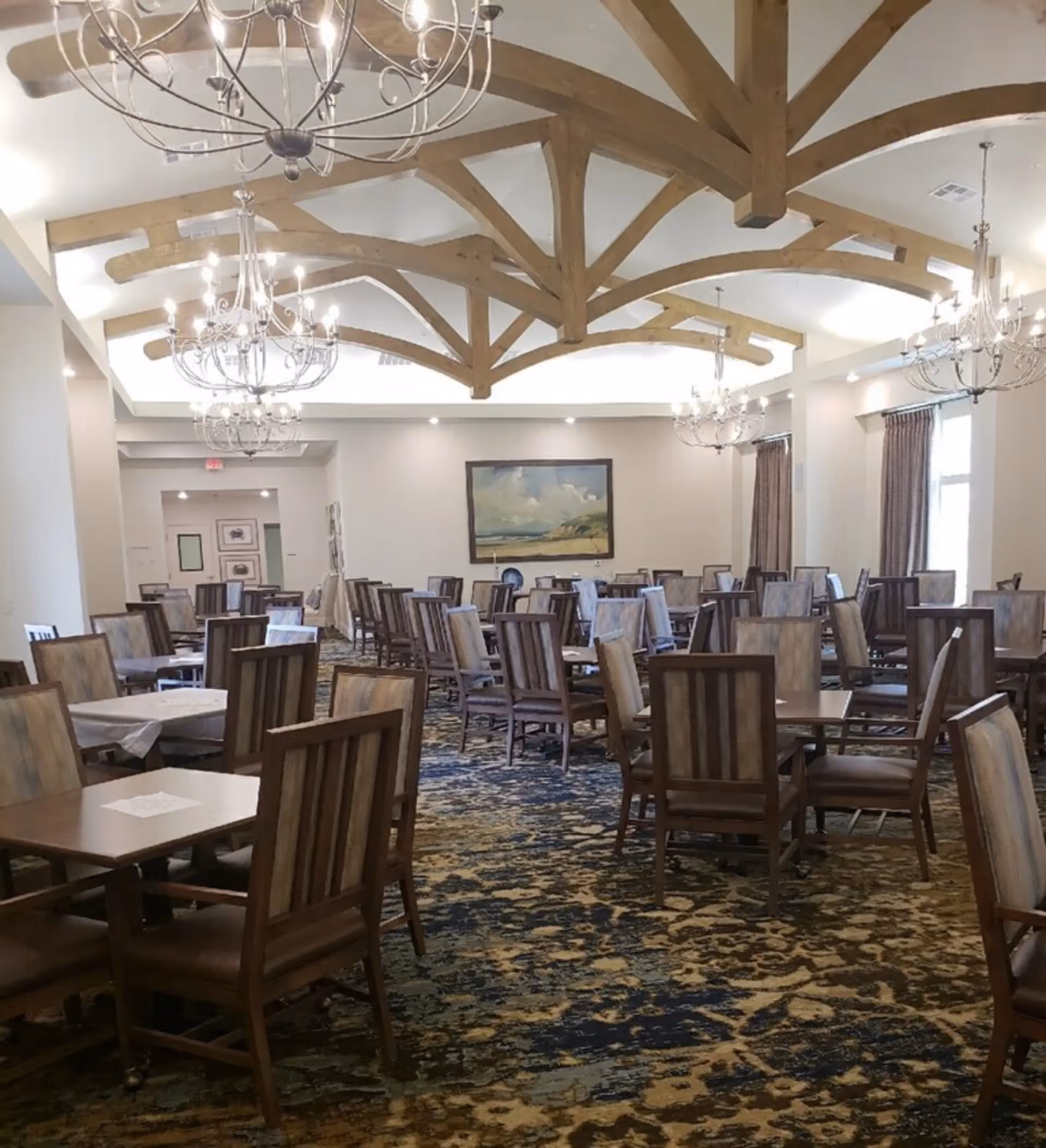 A spacious dining room with multiple wooden tables and chairs arranged neatly. The ceiling features exposed wooden beams and several elegant chandeliers providing warm lighting. The room has large windows with curtains and a framed landscape painting on the far wall. The carpet has a patterned design in shades of blue and beige.