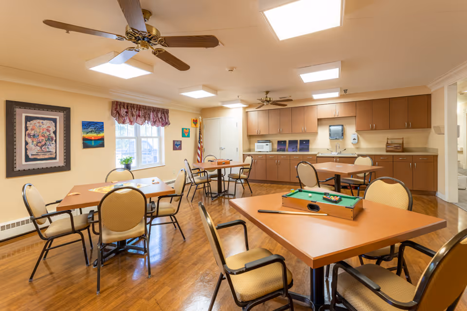 A well-lit common room with several tables and chairs arranged for group activities. The room features a ceiling fan, framed artwork on the walls, a window with a valance, and a kitchenette area with cabinets, a microwave, and a sink. One table has a small pool game set on it.