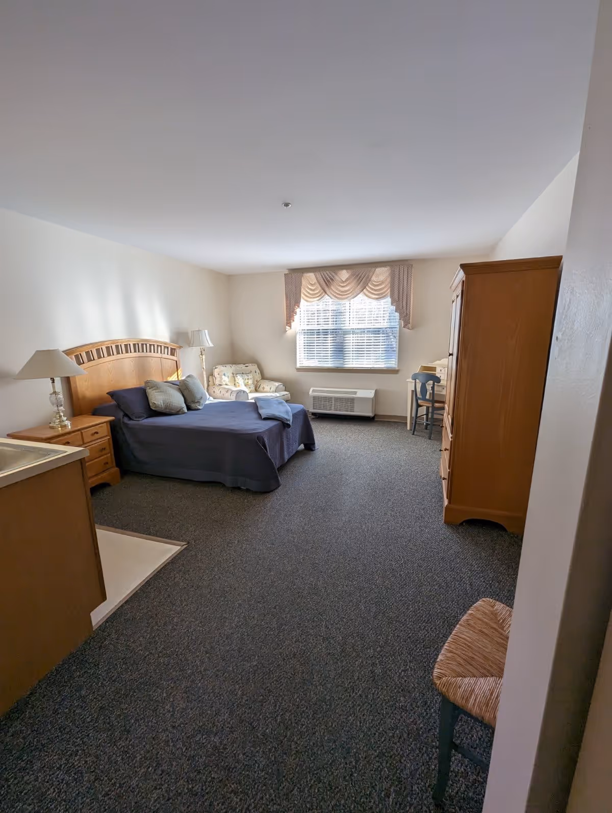 A senior living facility bedroom with a wooden bed frame, navy blue bedding, two pillows, a wooden nightstand with a lamp, a floral armchair near a window with blinds and a valance, a wooden armoire, a small desk with a chair, and a carpeted floor.
