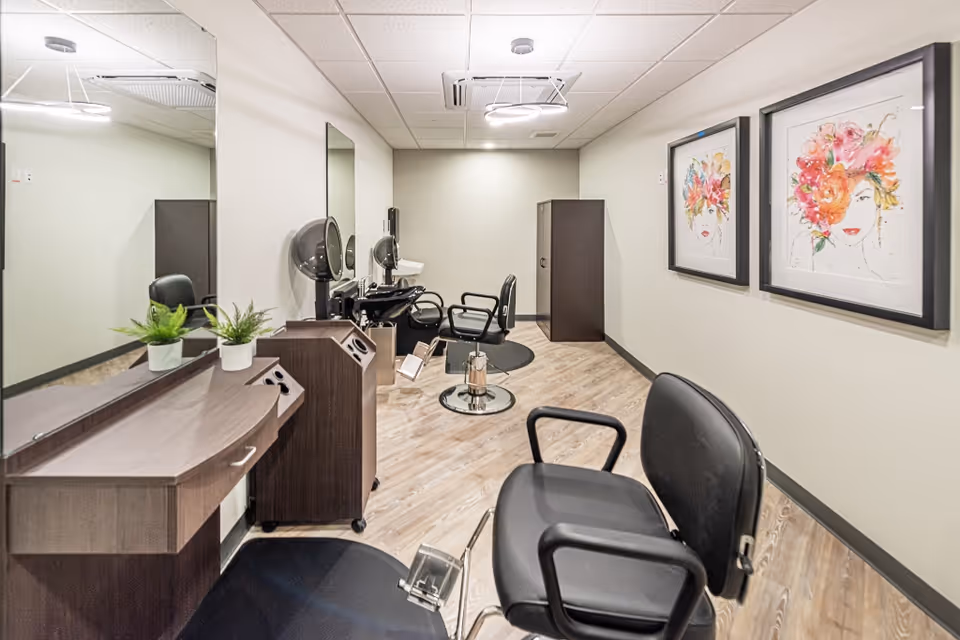 A bright salon room with styling chairs, mirrors, hairdryers, and framed floral artwork on the wall.