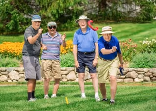 Four elderly men playing a lawn game on a grassy area with a stone wall and colorful flower beds in the background. They are outdoors on a sunny day, dressed casually in shorts, hats, and sunglasses, appearing to enjoy the activity together.