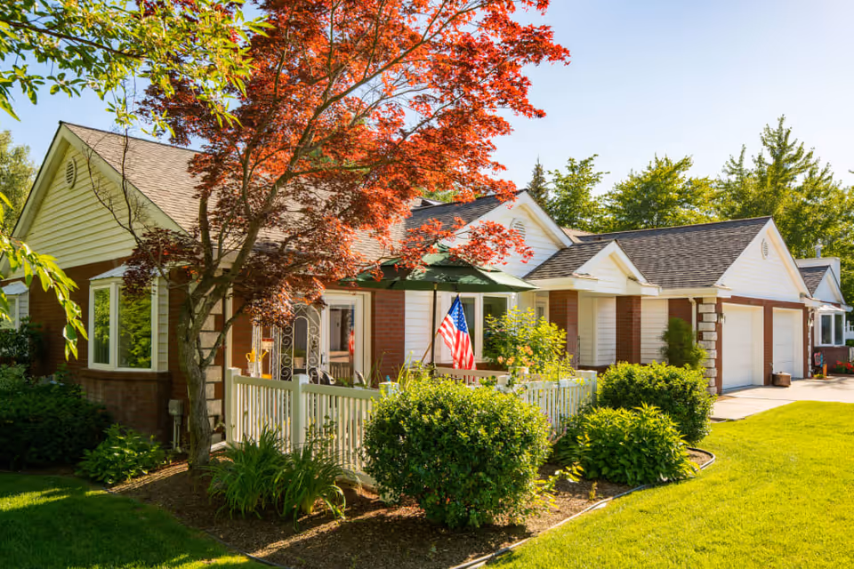 Front exterior of a ranch-style building with a porch, American flag, red-leaf tree, and manicured lawn.