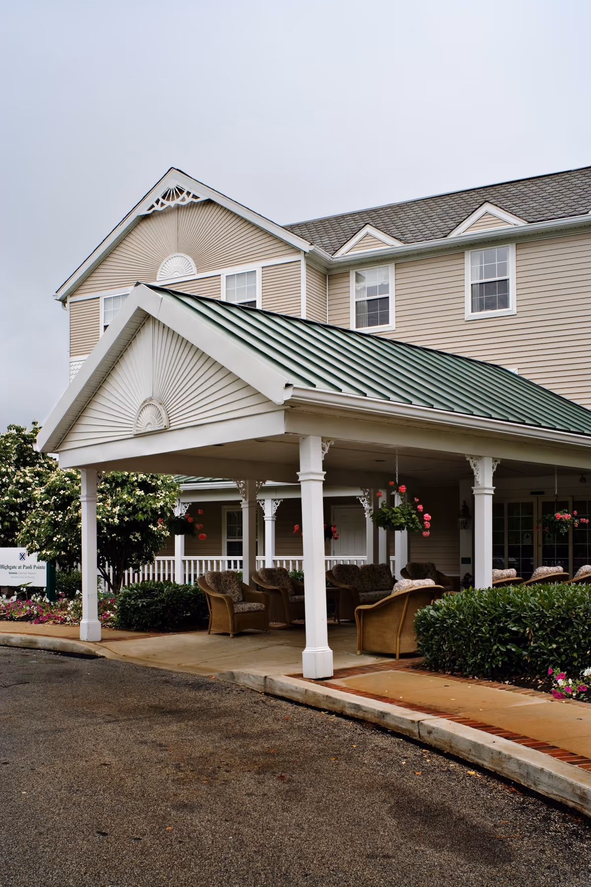 Exterior view of a senior living facility with a covered entrance supported by white columns. There are several cushioned wicker chairs arranged under the covered area, surrounded by bushes and hanging flower baskets. The building has beige siding and multiple windows, with a green metal roof over the entrance.