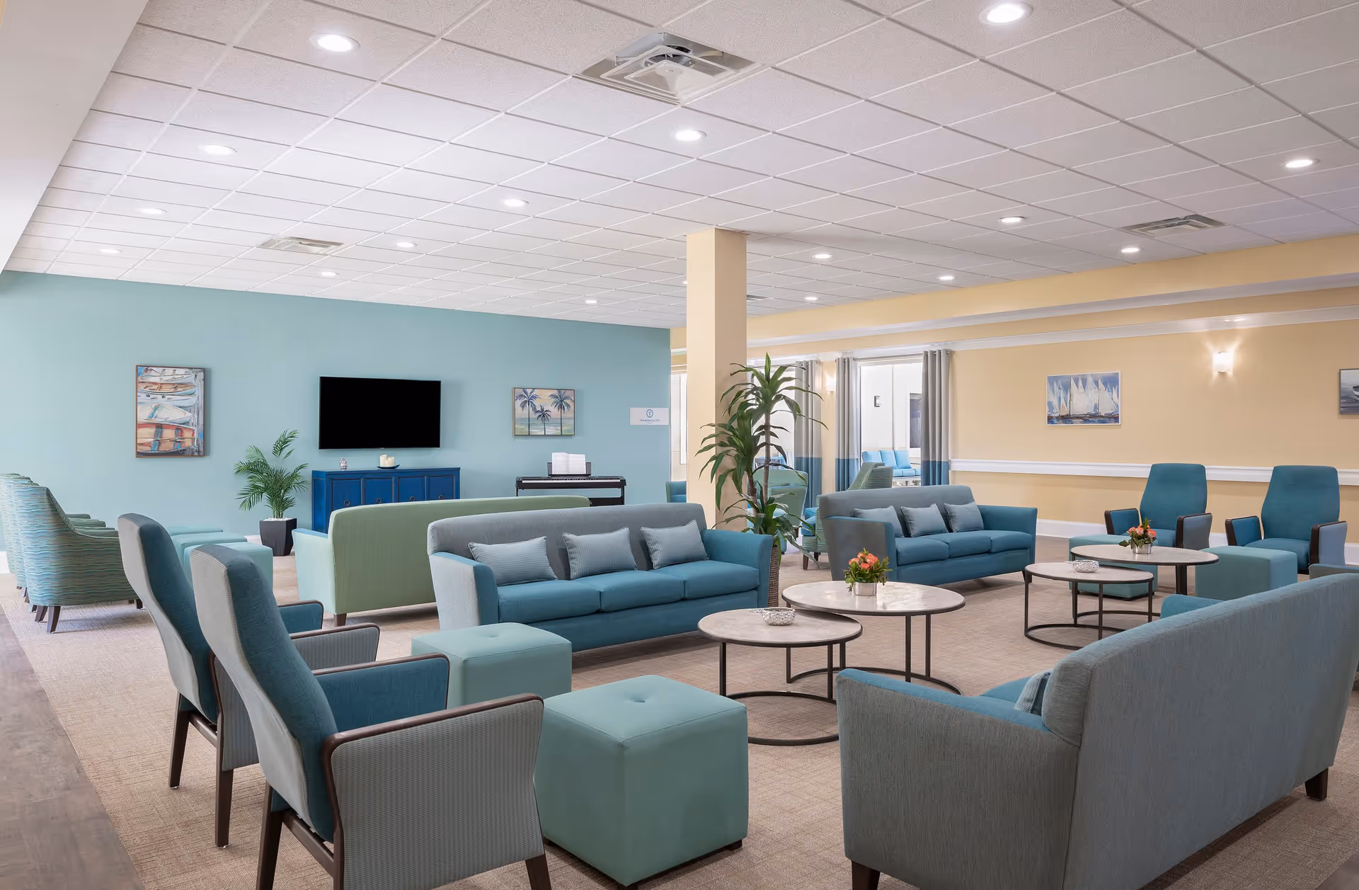 A spacious and well-lit senior living facility common area with multiple blue and teal sofas, armchairs, and ottomans arranged around round coffee tables. The walls are painted in soft pastel colors with framed artwork and a flat-screen TV mounted on one wall. There are plants and a piano in the background, creating a welcoming and comfortable atmosphere.