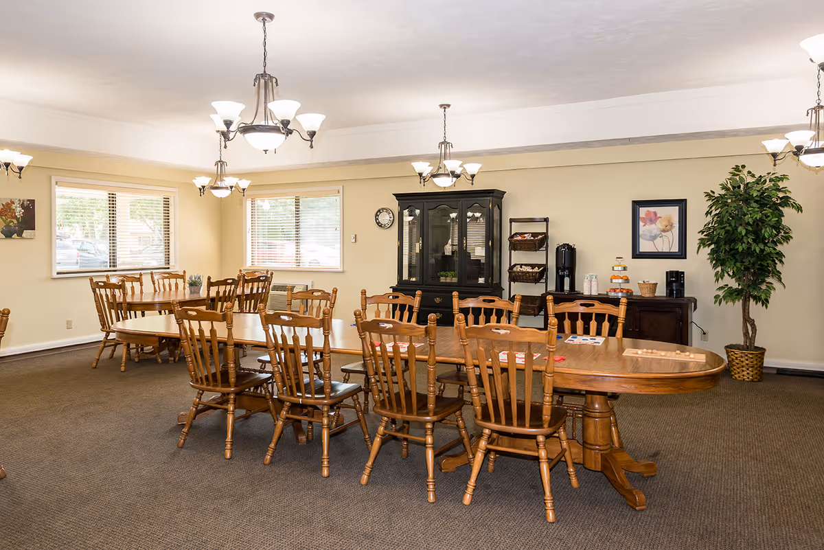 A spacious dining room with two long wooden tables surrounded by wooden chairs. The room has beige walls, carpeted floor, and multiple windows letting in natural light. There are three chandeliers hanging from the ceiling, a black china cabinet, a sideboard with coffee and snacks, a large potted plant, and framed artwork on the walls.