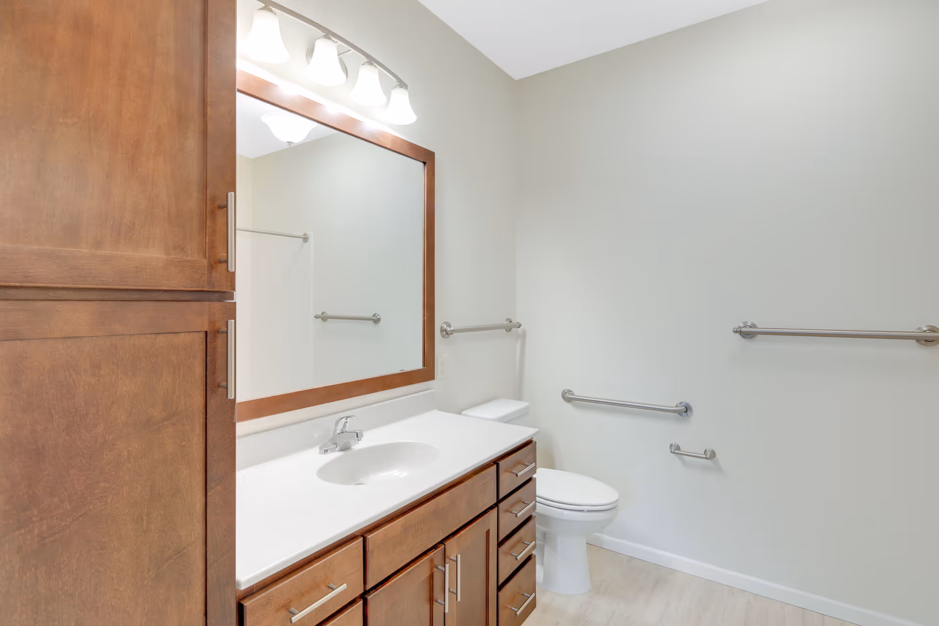 A clean and modern bathroom featuring a large wooden vanity with multiple drawers and a white countertop with an integrated sink. Above the sink is a large rectangular mirror framed in wood, illuminated by three overhead lights. The bathroom has light-colored walls and flooring, a white toilet, and several stainless steel grab bars mounted on the walls for accessibility.