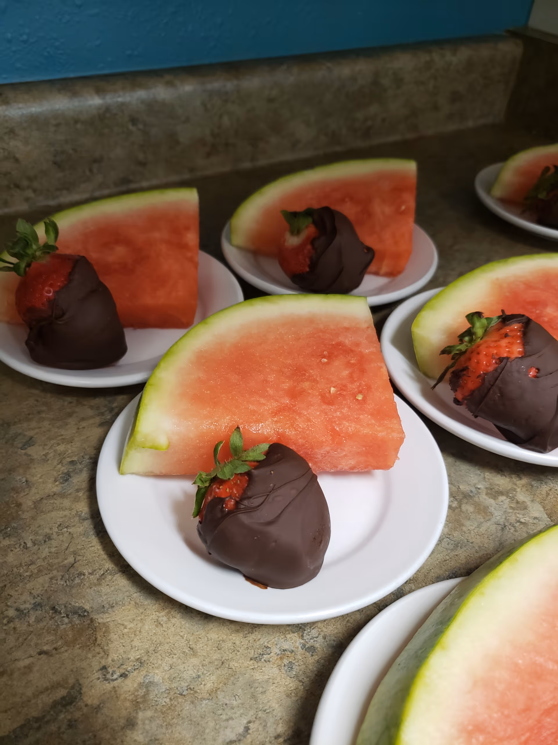 Several small white plates each holding a slice of watermelon and a chocolate-covered strawberry, arranged on a countertop.