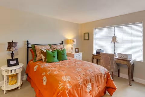 A cozy bedroom with an orange floral comforter, green pillows on a metal-framed bed, bedside tables with lamps, and a desk by a window with blinds.
