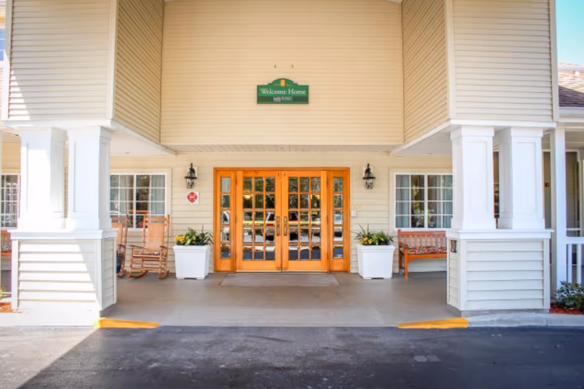 Entrance of a senior living facility with double wooden doors, two large white pillars, two potted plants, rocking chairs, and a bench on the porch. A green sign above the doors reads 'Welcome Home'.