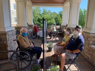 Four elderly individuals sitting outside on a covered porch area, separated by a clear plastic divider. Two people are in wheelchairs on the left side, and two people are seated on chairs on the right side. All are wearing face masks. The porch has stone pillars and overlooks greenery in the background.