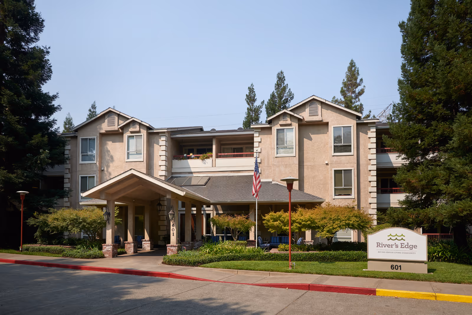 Exterior front view of River's Edge senior living community building with a covered entrance, American flag, trees, and a sign displaying the facility name and address number 601.