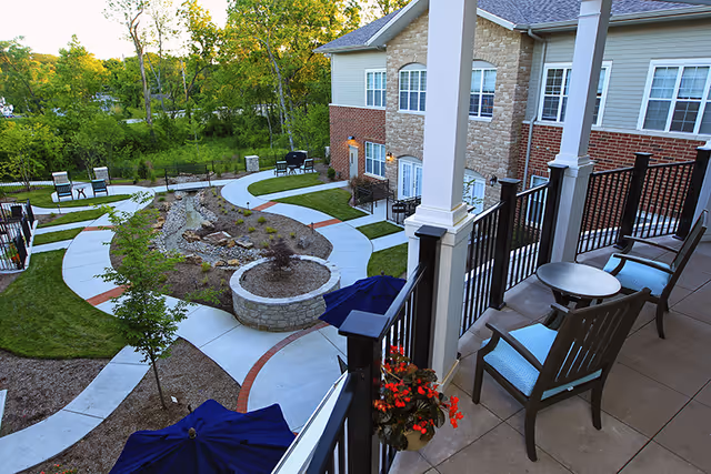 View from a balcony overlooking a landscaped outdoor courtyard with winding concrete pathways, small trees, benches, and patio tables with umbrellas. The building has a brick and stone exterior with multiple windows and doors leading to the courtyard.