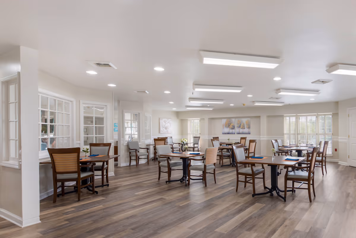 A bright and spacious dining room with multiple wooden tables and chairs arranged neatly on a wood-patterned floor. The room features large windows with white blinds, soft wall art, and recessed ceiling lights providing ample illumination.