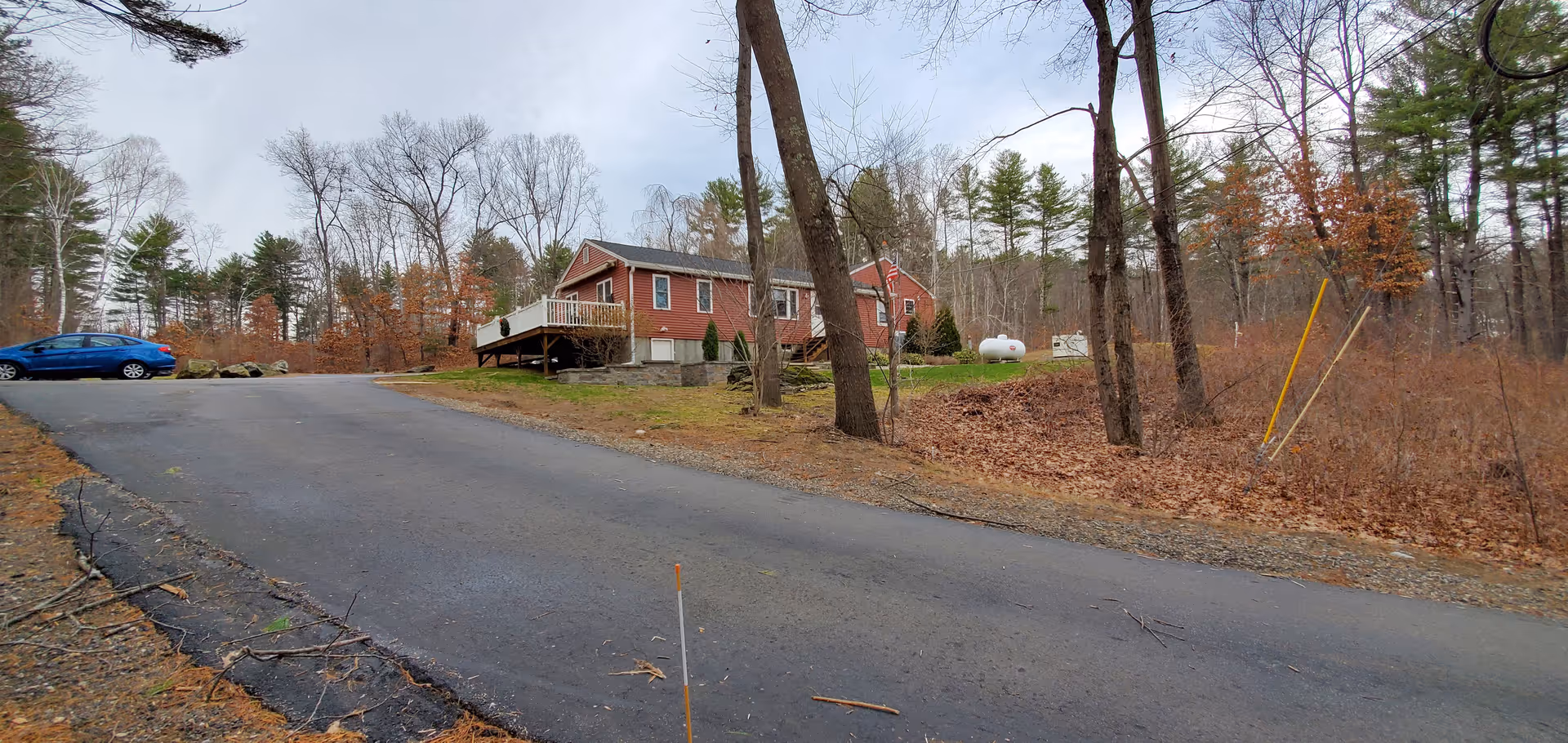 A paved driveway leading up to a red single-story house with a small deck, surrounded by leafless trees and dry foliage in a wooded area under a cloudy sky. A blue car is parked on the left side near the house.