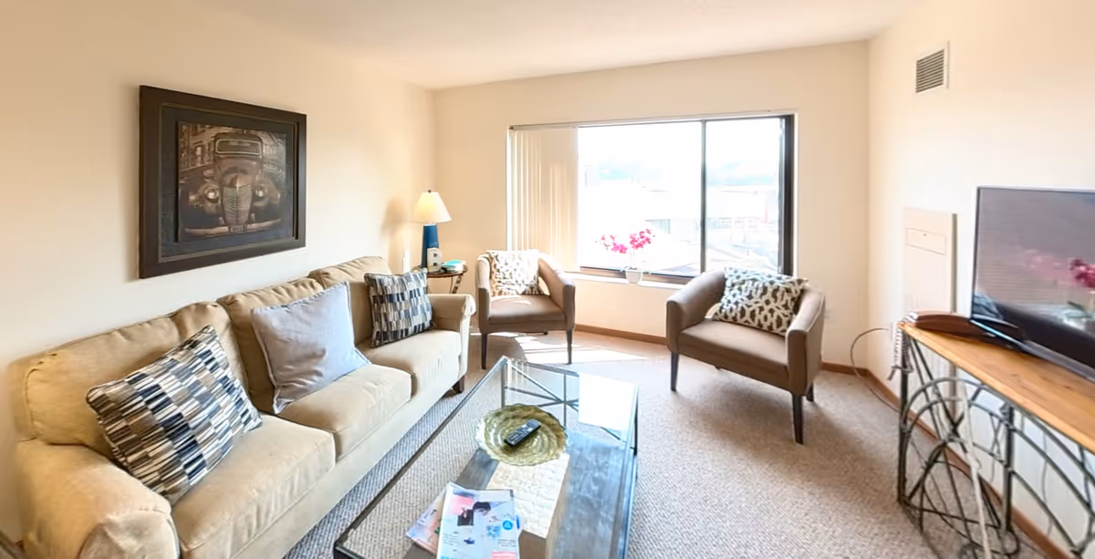 A cozy living room with a beige sofa adorned with three patterned pillows, two brown armchairs with patterned cushions, a glass coffee table with magazines and a decorative bowl, a floor lamp, a framed vintage car picture on the wall, and a large window with vertical blinds letting in natural light. A TV is placed on a wooden and metal stand on the right side of the room.
