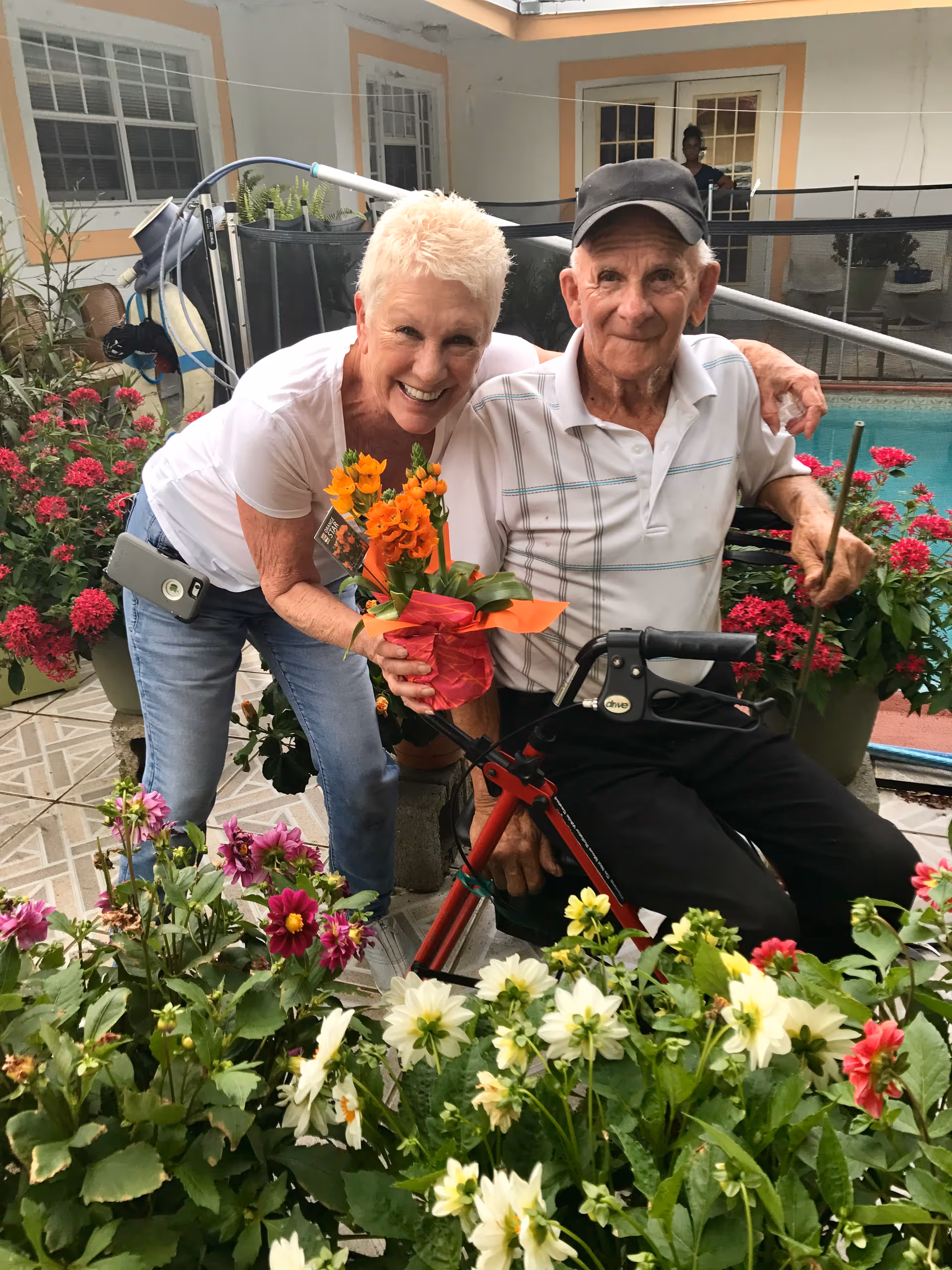 An elderly man sitting on a walker and a woman with short blonde hair smiling and holding a potted orange flower, surrounded by various colorful flowers in an outdoor patio area with a swimming pool in the background.