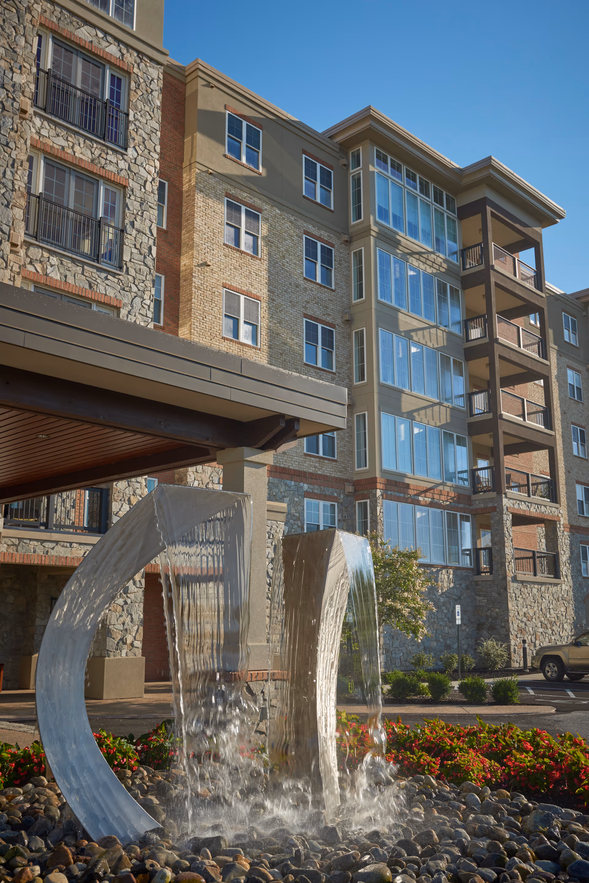 Exterior view of a multi-story senior living facility building with stone and brick facade, featuring large windows and balconies. In the foreground, there is a modern water fountain with two curved metal structures with water flowing down into a bed of rocks, surrounded by red flowers and greenery under a clear blue sky.