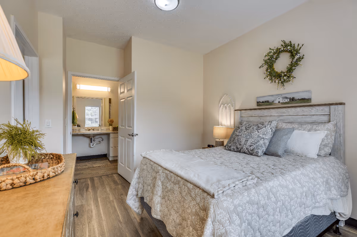 A cozy bedroom with a large bed covered in a light gray and white patterned quilt and multiple pillows. Above the bed is a decorative wreath and a small landscape painting. To the left, there is a wooden dresser with a lamp and a woven basket holding a small plant. The door to an adjoining bathroom is open, showing a sink with a large mirror and a window in the bathroom. The room has wood flooring and neutral-colored walls.