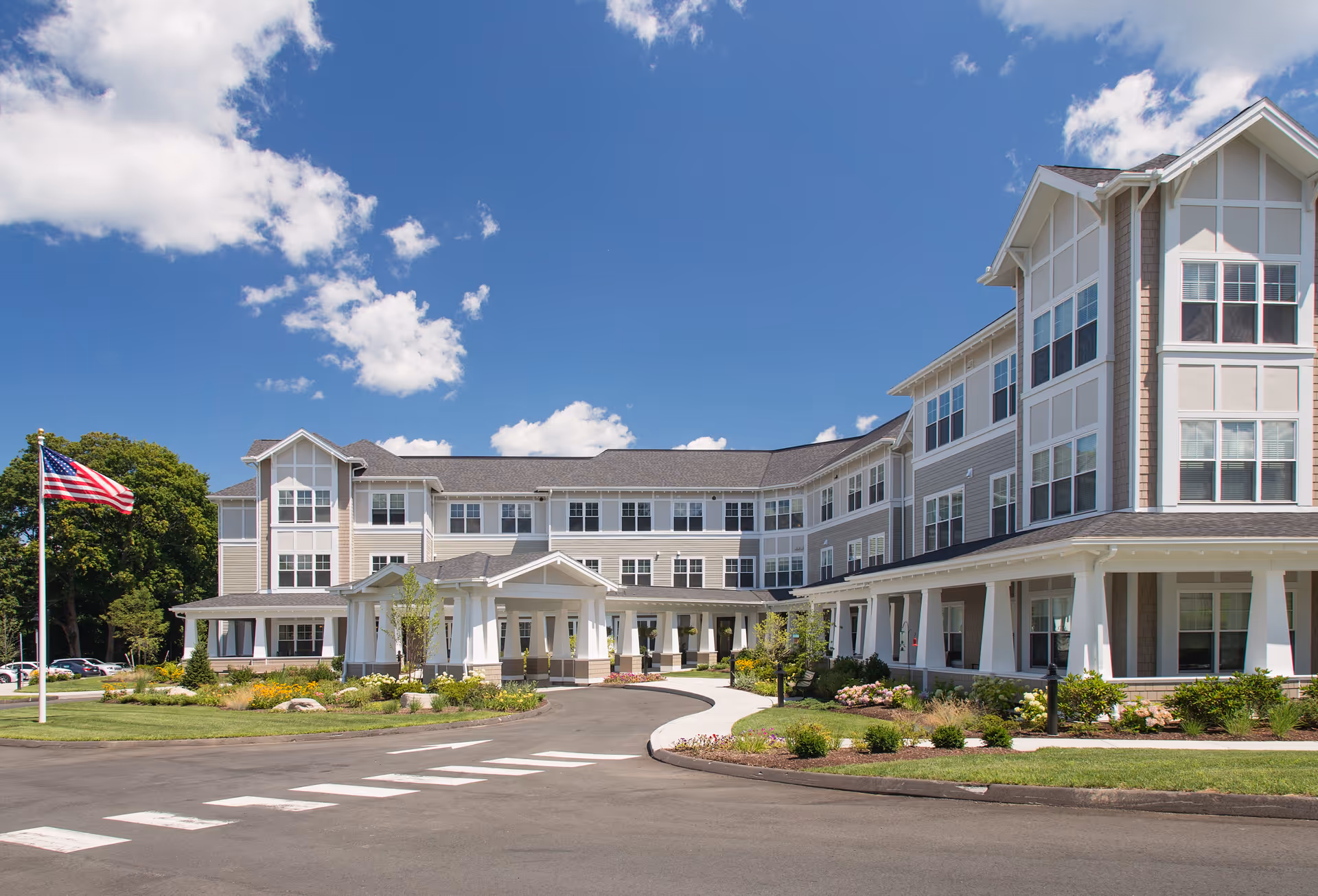 Exterior view of a large, modern senior living facility building under a bright blue sky with scattered clouds. The building has multiple stories with many windows and a covered entrance. There is a well-maintained driveway, landscaped greenery, and an American flag on a flagpole in front.