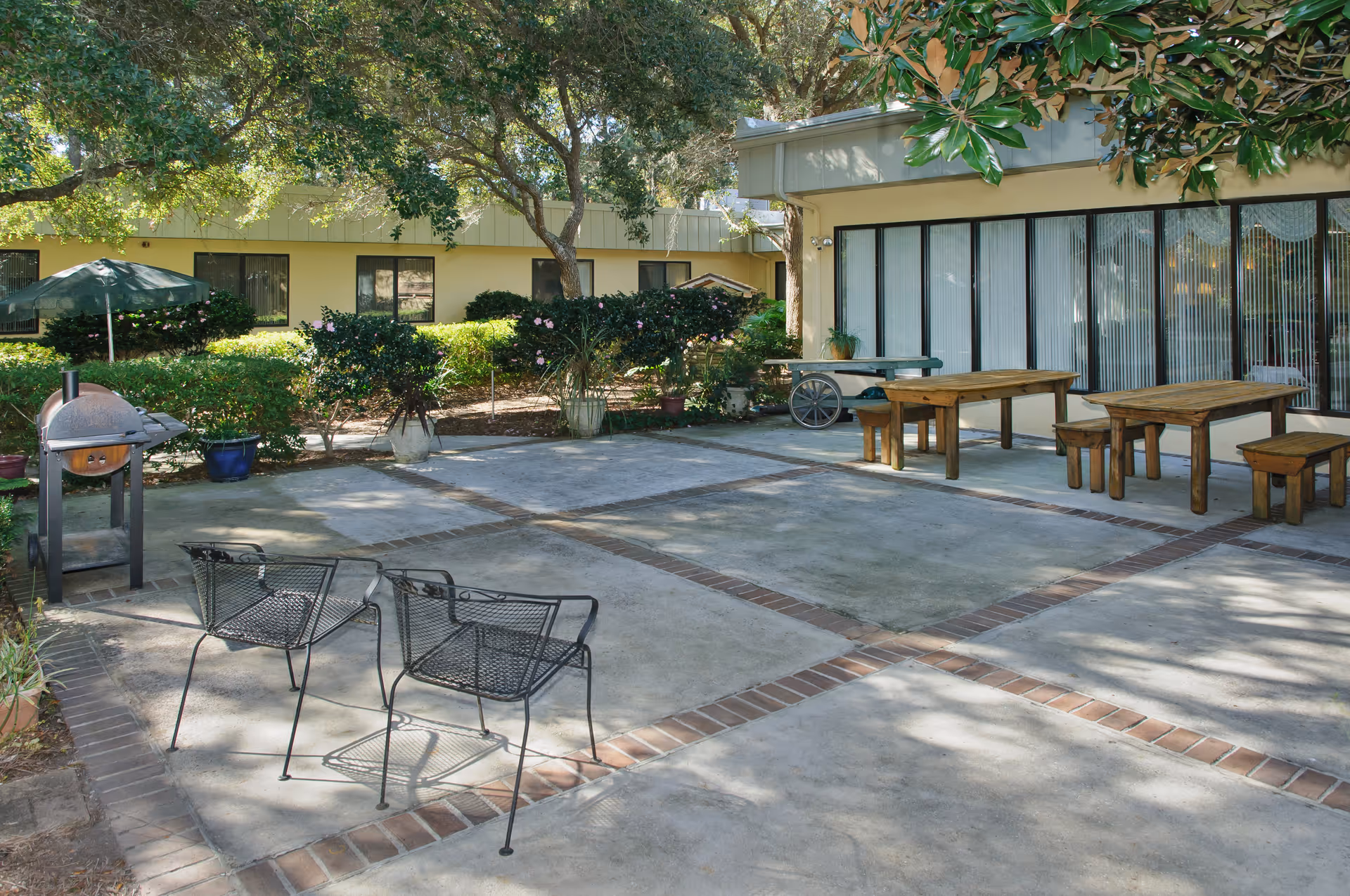 Outdoor patio area with concrete flooring bordered by bricks, featuring two black metal chairs, wooden picnic tables with benches, a small barbecue grill, and surrounded by greenery and trees. The building with large windows and vertical blinds is visible in the background.