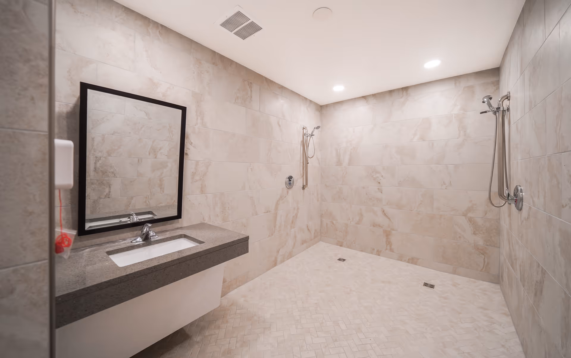 A spacious, modern bathroom with beige marble tiled walls and floor. The room features two wall-mounted shower heads with handheld attachments on the right side and a large rectangular mirror above a gray countertop with a built-in sink on the left side. The ceiling has recessed lighting and a ventilation vent.