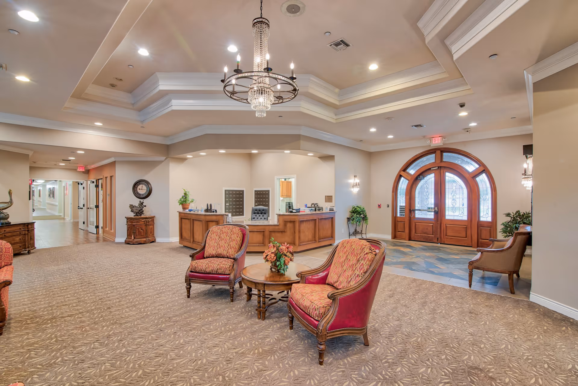 Spacious and well-lit assisted living community lobby with a chandelier hanging from a tray ceiling, two upholstered armchairs and a round wooden table with a floral arrangement in the center, a wooden reception desk in the background, and large arched wooden double doors with glass panels on the right side.