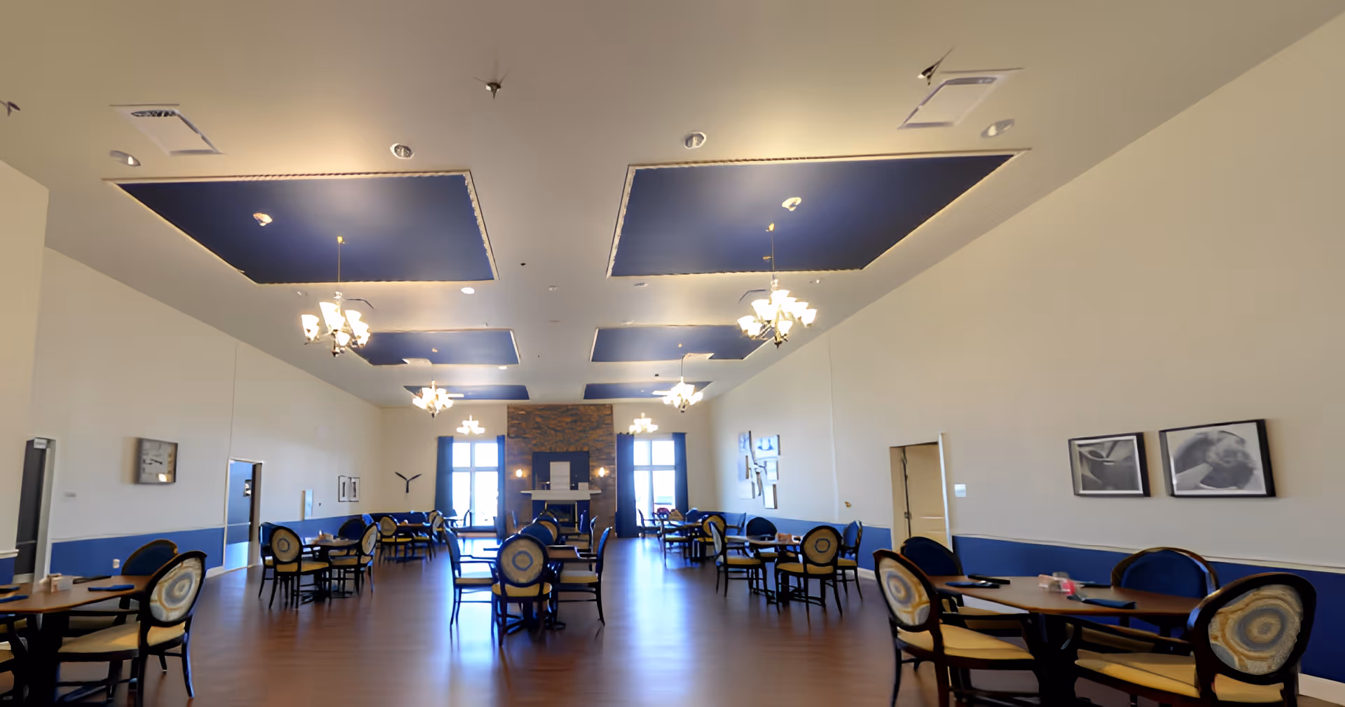 A spacious dining room with multiple round tables and chairs arranged neatly. The ceiling features blue recessed panels with hanging chandeliers. The room has large windows at the far end with blue curtains and a fireplace in the center. The walls are decorated with framed pictures and the floor is wooden.