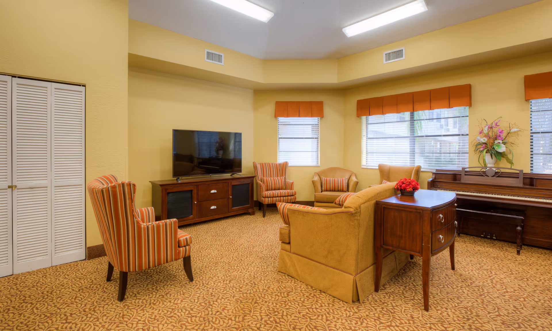 A cozy living room with yellow walls and patterned carpet. The room features a flat-screen TV on a wooden stand, a piano with a floral arrangement on top, a yellow sofa, and several armchairs with striped upholstery. Windows with orange valances let in natural light.