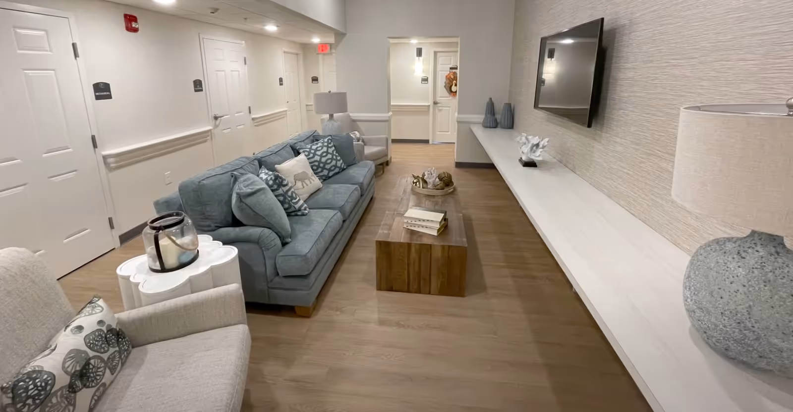 Lounge area in a senior living facility featuring a blue sofa and armchairs arranged around a wooden coffee table, a wall-mounted TV, and a long shelf along the wall.