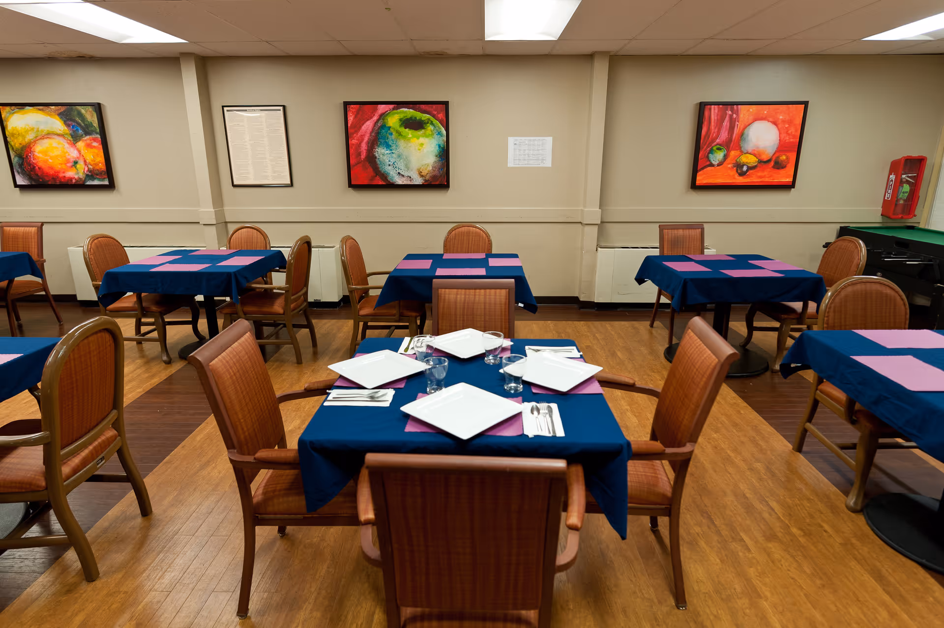 A dining room with several tables covered in blue tablecloths and pink placemats. Each table is surrounded by wooden chairs with cushioned seats. The table in the foreground is set with white square plates, glasses, and silverware. The walls are decorated with colorful fruit-themed paintings, and there is a fire extinguisher mounted on the right wall.