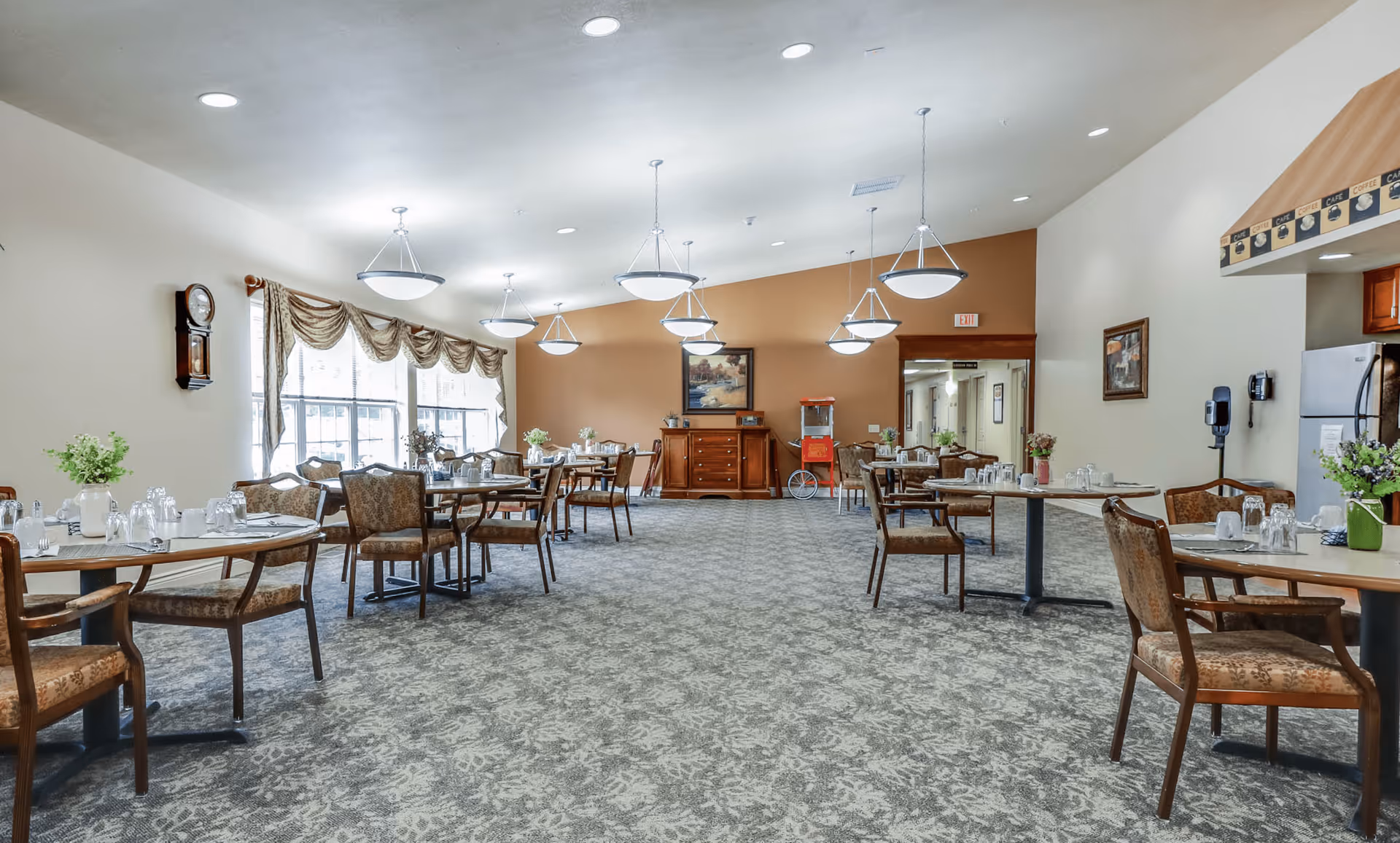 A spacious dining room in an assisted living facility with multiple tables and chairs arranged neatly. The room has large windows with draped curtains allowing natural light to enter. There are several hanging light fixtures on the ceiling, a wall clock, and a popcorn machine near the back wall. The carpet has a patterned design, and there are decorative plants and framed artwork on the walls.